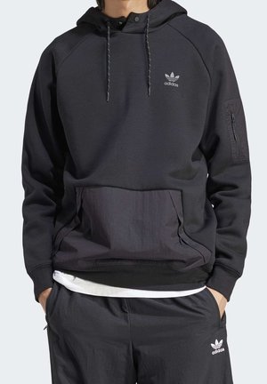 Homme portant un sweat à capuche Adidas noir avec une poche ventrale et un pantalon Adidas noir assorti, les mains dans les poches, debout devant un fond neutre.