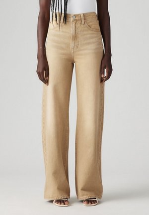 Vrouw draagt beige high-waisted wijde jeans en witte sandalen, staand tegen een effen lichtgrijze achtergrond.
