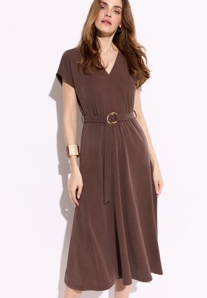 Kobieta ubrana w brązową sukienkę midi z dekoltem w serek, krótkimi rękawami oraz dopasowanym paskiem z złotą klamrą, uzupełniona szeroką, złotą bransoletą.