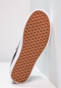 Semelle de chaussure avec motif gaufré en caoutchouc brun sur une basket blanche et noire reposant contre un mur et un sol blancs.