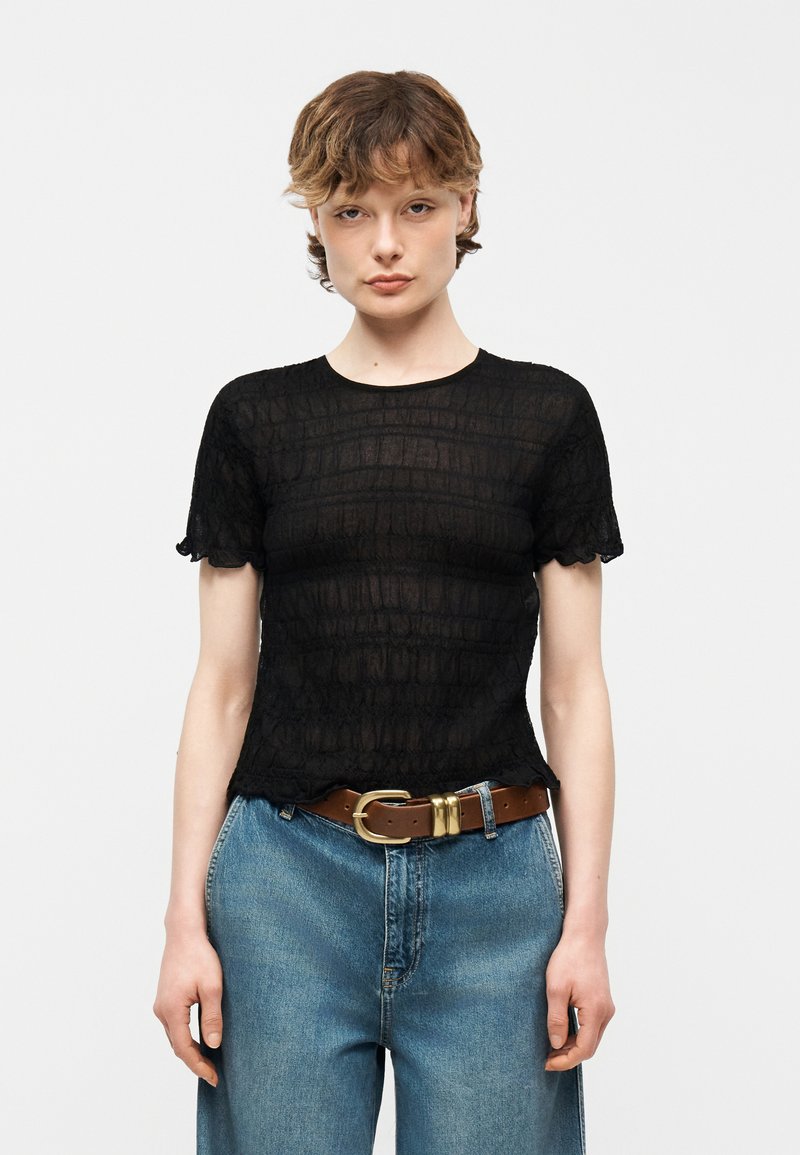 Νεαρός με κοντά μαλλιά, φορώντας μαύρο υφασμάτινο μπλουζάκι με κοντά μανίκια, μπλε τζιν και καφέ ζώνη, στέκεται μπροστά από ένα απλό φόντο.