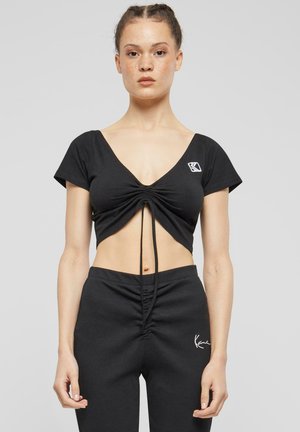 T-shirt noir court en tissu doux, avec un décolleté en V profond, un détail froncé à l'avant et des manches courtes. Petit logo sur la poitrine.