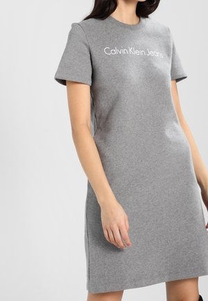 Kvinde iført en kortærmet, knælængde grå kjole med "Calvin Klein Jeans"-logo på brystet, stående mod en ensfarvet baggrund.