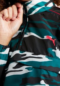 Svart, teal och vit kamouflagemönstrad jacka med dragkedja i halsen och röd Puma-logotyp. Materialet verkar lätt och slätt.