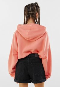 Sudadera coral de gran tamaño con una capucha grande, textura suave y hombros caídos, combinada con pantalones cortos de mezclilla negros de tiro alto.