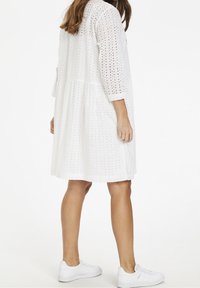 Weißes Broderie-Dress mit Dreiviertelärmeln, einem ausgestellten Saum und einem gestuften Design. Kombiniert mit weißen Sneakers. Sanfte Stofftextur.
