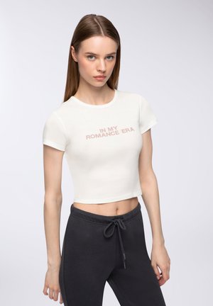 T-shirt bianco corto con scollo rotondo e maniche corte, con il testo "IN MY ROMANCE ERA" in rosa chiaro. Pantaloni della tuta grigio scuro.