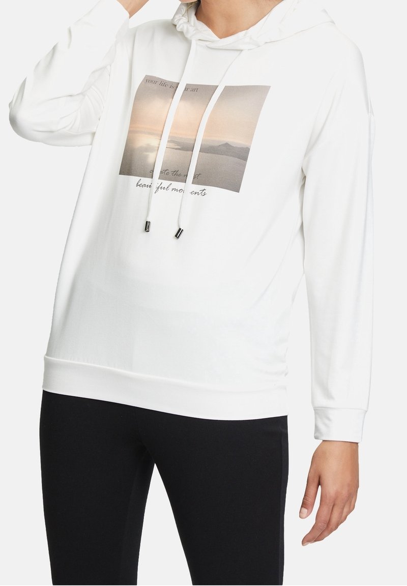 Personne portant un sweat à capuche blanc avec cordons et un graphique en trois panneaux représentant un coucher de soleil montrant de l'eau et des montagnes, associé à un pantalon noir.
