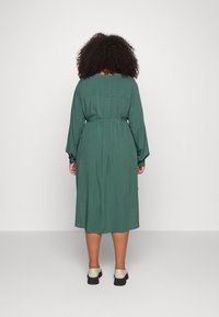 Zelené šaty střední délky s dlouhými rukávy, nařaseným pasem a volným střihem. Hladká tkanina, detail na zadním díle, doplněné světle zbarvenou obuví.