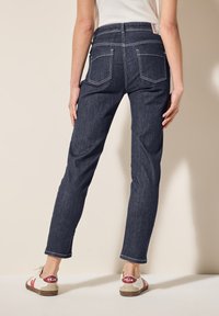 Jeans de mezclilla azul oscuro con un diseño de pierna recta, con costuras blancas y dos bolsillos traseros. Llevados con zapatillas blancas y una blusa clara.