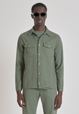 Olive green, button-up jacket with two chest pockets, contrasting stitching, and a smooth texture. Worn with matching pants and sunglasses.