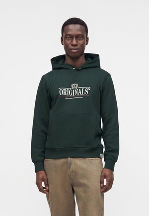 Homme portant un sweat à capuche vert foncé avec le texte "ORIGINALS", et un pantalon marron clair, debout devant un fond clair uni.