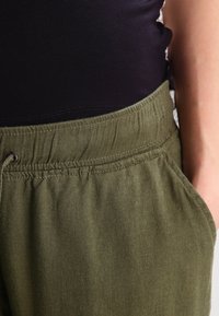 Olive green elastic waistband pants with side pockets, made from a soft, textured fabric. The waistband includes a drawstring detail.