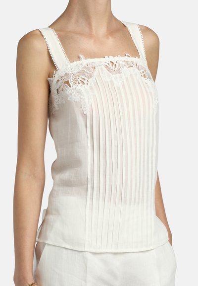 Haut blanc sans manches avec plis verticaux et bordure en dentelle autour de l'encolure, porté avec un pantalon blanc.