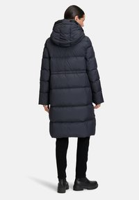 Giacca piumino grigia scura con cappuccio, caratterizzata da un design trapuntato e una vestibilità oversize, che arriva a metà polpaccio. Indossata sopra pantaloni neri e stivali.