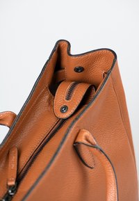Braune Lederhandtasche mit griffiger Textur, akzentuiert durch einen schwarzen Reißverschluss und einen Druckknopfverschluss. Verfügt über robuste Griffe und ein minimalistisches Design.
