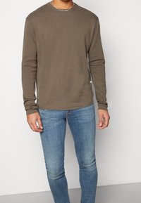 Hombre con un suéter de manga larga verde oliva y jeans ajustados de color azul claro de pie frente a un fondo blanco liso.