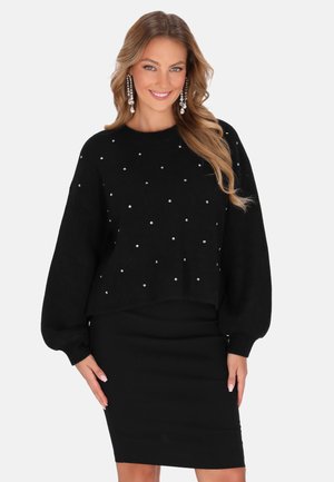 Kvinde med langt bølget hår iført en sort sweater med små hvide perler og en tætsiddende sort nederdel, smilende med hængende perleøreringe.