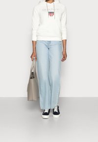 Weißer Kapuzenpullover mit Logo und Schilddetail, hellblaue Jeans mit weitem Bein, marineblaue Sneakers mit weißen Sohlen und eine graue Tragetasche.