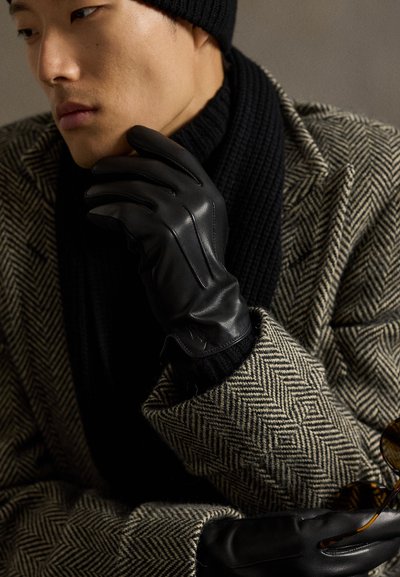 Svarte lærhansker med en glatt tekstur og detaljert søm, båret på en hånd mot en fiskebensmønstret frakk i dempede toner.