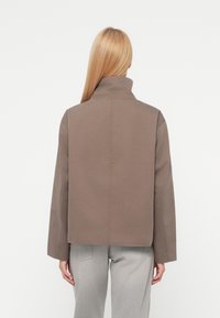 Personne blonde portant une veste marron à col montant et un jean gris clair, debout de dos face à la caméra sur un fond blanc uni.