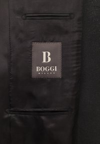 Juodas audinys su lygiu paviršiumi ir pritaikytu siluetu; turi siuvinėtą etikėtę su "B BOGGI MILANO" šviesiai pilka spalva.