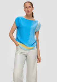 Blaue und weiße Kurzarmbluse mit gelben Akzenten; glatte Textur, lockerer Schnitt, getragen mit hellen, weit geschnittenen Hosen.