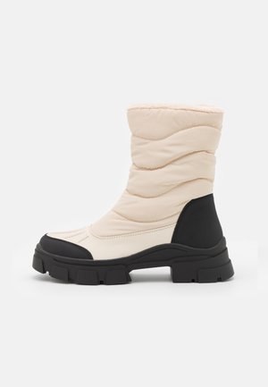 Botte d’hiver mi-mollet avec dessus matelassé crème et semelle et talon en caoutchouc noir épais, conçue pour temps froid et usage extérieur.
