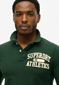 Groene polo shirt van katoen met een kraag, voorzien van crèmekleurige geborduurde tekst "SUPERDRY ATHLETICS" en knoopdetails op de knoopsluiting.