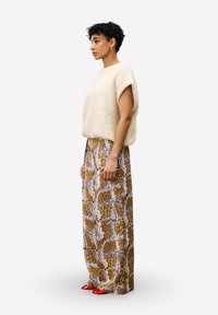 Femme aux cheveux courts et bouclés, portant un pull sans manches crème, un pantalon large à motifs et des chaussures rouges à bout pointu, se tenant de profil.
