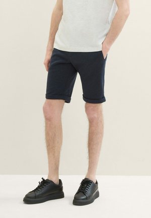 Shorts en coton navy avec ourlets retournés, poches latérales, associés à des baskets en cuir noir avec une semelle épaisse et des lacets.