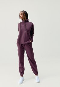 Lila atletisk outfit med en höghalsskjorta i lång ärm och tapered byxor med elastiska manchetter, tillsammans med vita slip-on skor.