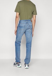 Vaqueros de mezclilla azul con un corte recto, cinco bolsillos y costuras en contraste, combinados con una camiseta verde y zapatillas deportivas blancas.