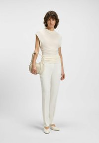 Vaalean kermaisin blouse, jossa on rypytetty pinta, yhdistettynä suoriin kermaisiin housuihin ja soveltuviin kärkikenkkiin sekä pieneen pyöreään käsilaukkuun.