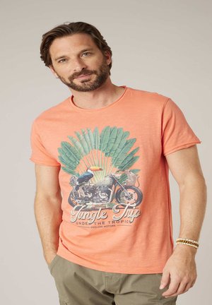 Homme portant un t-shirt corail orné de feuilles tropicales, d'un toucan, d'une moto et d'une souris, associé à un pantalon cargo olive et des bracelets perlés.