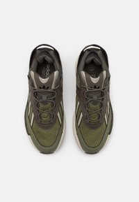 Sneakers med ett nät tyg i grönt och brunt, rundad tå, snörning och diskreta vita ränder längs sidorna.