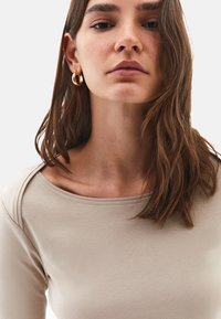 Junge Frau mit braunen Haaren trägt goldene Creolen und ein beiges Oberteil, schaut geradeaus vor einem schlichten hellen Hintergrund.