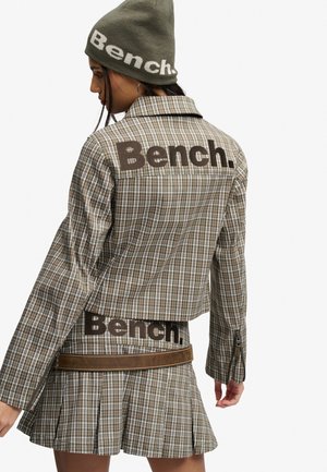 Kobieta ubrana w brązowo-białą kratowaną kurtkę, plisowaną spódnicę i oliwkową czapkę, wszystko z logo "Bench.", odwrócona tyłem, z jedną ręką opuszczoną.