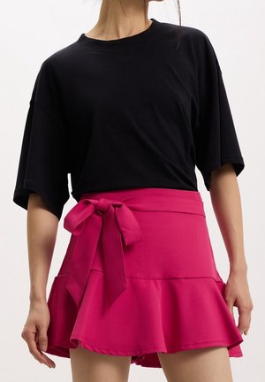 Femme portant un haut noir ample à manches courtes et une jupe rose vif avec un grand nœud sur le côté et un bas évasé, sur fond blanc.