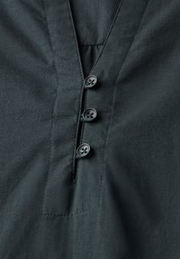 Dunkelgrünes Shirt mit V-Ausschnitt, ausgestattet mit vier schwarzen Knöpfen und einem gerafften Detail am Ausschnitt. Glatte Stoffqualität mit subtiler Textur.