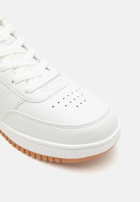 Fehér bőr sportcipő, lekerekített orral, struktúrált felülettel, perforált részletekkel és kontrasztos barna gumitalppal.