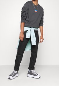 Muž ve tmavě šedé mikině značky Levi's, černých vykasaných kalhotách, šedých vysokých platformových teniskách a světle modré košili uvázané kolem pasu.