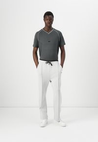 A young man stands confidently, wearing a gray v-neck t-shirt and light gray trousers, with hands in pockets, set against a plain background.