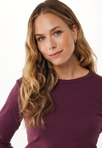 Junge Frau mit langen welligen blonden Haaren, die ein lila geripptes Oberteil trägt und sanft gegen einen schlichten weißen Hintergrund lächelt.