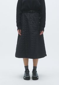 Saia preta acolchoada com um design de comprimento médio, apresentando um cinto na cintura. Combinada com sapatos pretos e meias cinzentas. Textura suave.