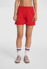 Röda idrottsshorts med elastisk midja, med grå detaljer på sidorna. Kombineras med vita strumpor och beige sportskor.