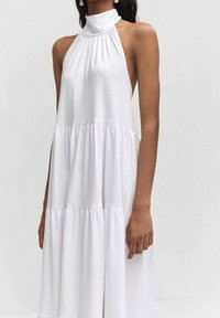Biała sukienka na ramiączkach wykonana z miękkiego materiału. Posiada marszczone detale przy dekolcie oraz warstwowy krój. Sięga do długości w połowie łydki.