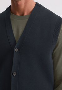 Marineblaue Strickweste mit V-Ausschnitt und vier Knöpfen, über einem hellolivgrünen Shirt getragen, das gerippte Akzente enthält.