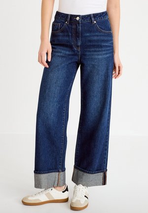 Donna che indossa jeans blu scuro a gamba larga con risvolti e sneakers bianche con accenti neri e beige, in piedi davanti a uno sfondo bianco semplice.