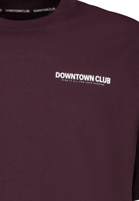 Burgundy long-sleeve t-shirt made of smooth fabric. Features white text reading "DOWNTOWN CLUB" and a slogan near the neckline.